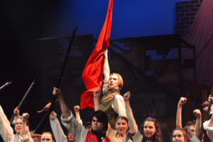 A revolutionary, played by David Nielsen (12), holds up a red flag, one of the symbols for the revolution, The students built a barricade and tried to fight the French army, but the battle ended in a painful defeat where all men except Marius and Valjean died.