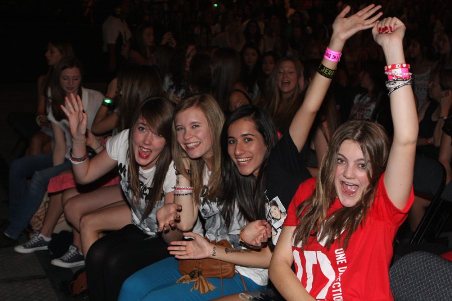 Fangirls smile at a One Direction concert, dressed in the boybands merchandise. The girls excitedly wave at the camera during the event. Fangirls receive significant criticism and are often degraded for their likes. Its time we stop associating female joy with shame, and learn to celebrate excitement.