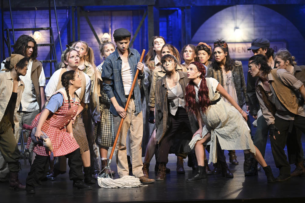 Urinetown cast members gather around the main character, Bobby Strong (Luke Dunphy). Bobby’s father was just taken away by the police for peeing illegally. This spurs the idea to lead a revolution against a monopolistic corporation known as Urine Good Company that regulates water consumption and restroom usage.