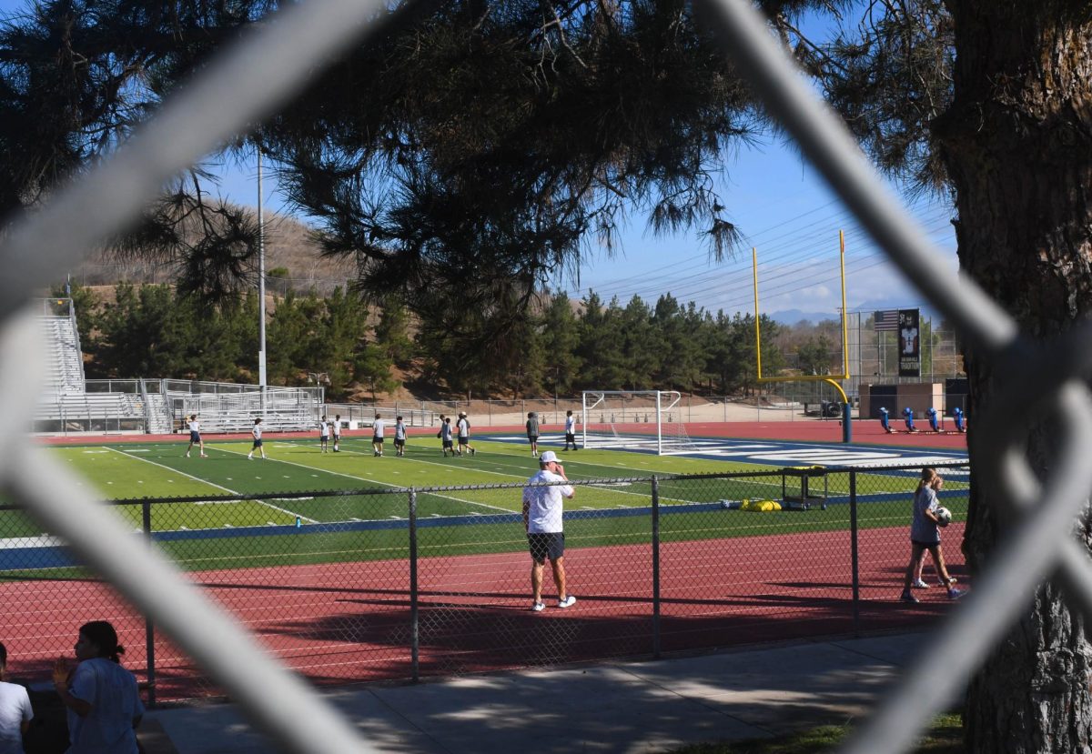 Students at PE play soccer on the football field while Coach Cuculic supervises. Other students take a break in the shade or walk around for exercise during the first period.