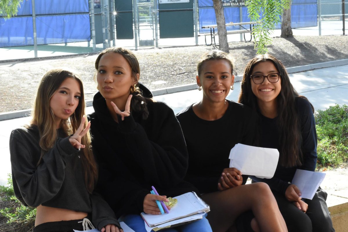 Spanish 1 rehearses scripts outside of E building to help practice Spanish dialogue. Students Imogen Tinney (9), Saphira Millers (10), Bella Sanchez (9), and Janice Sterling (9) are pictured smiling.